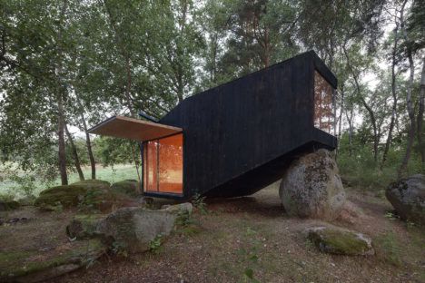 A modern looking black house in a forest propped up against a large rock. 