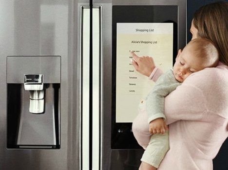 A woman holding a baby and using the touch screen on a modern refrigerator.