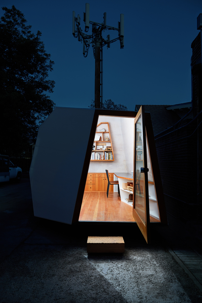 The Lighthouse backyard office pod at night, with the door open and light on inside. 