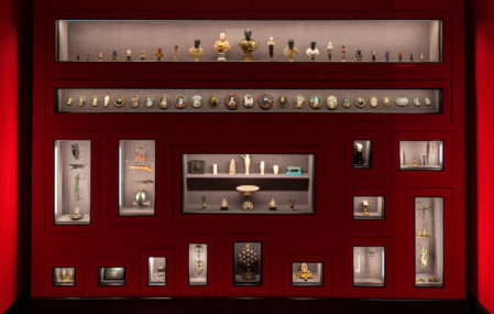 A lush red wall lined with display cases full of historical artifacts, including busts, portraits, and statues. Curated by Wes Anderson and partner Juman Malouf as part of their "Spitzmaus Mummy in a Coffin and Other Treasures" exhibit.
