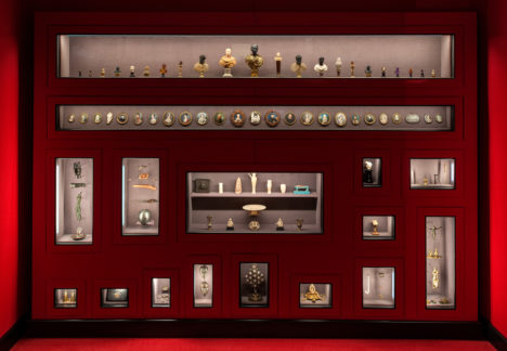 A lush red wall lined with display cases full of historical artifacts, including busts, portraits, and statues. Curated by Wes Anderson and partner Juman Malouf as part of their "Spitzmaus Mummy in a Coffin and Other Treasures" exhibit.
