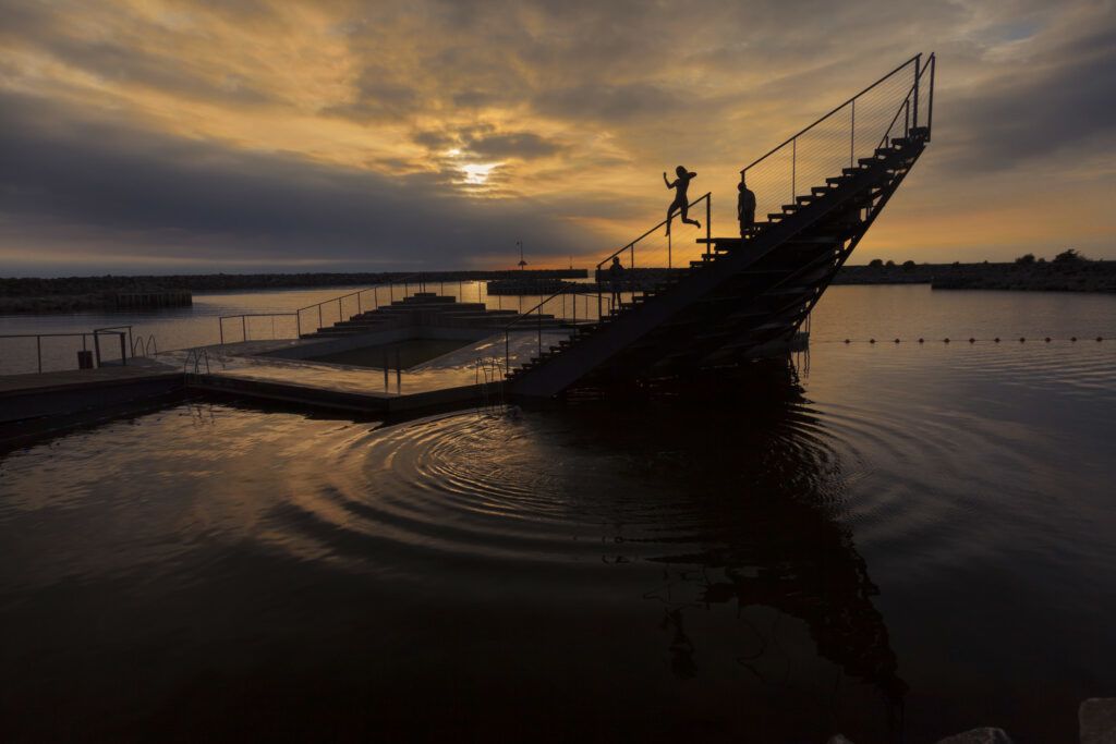 People jump off the edge of the Hasle Harbour Bath as the sun sets over the seas.